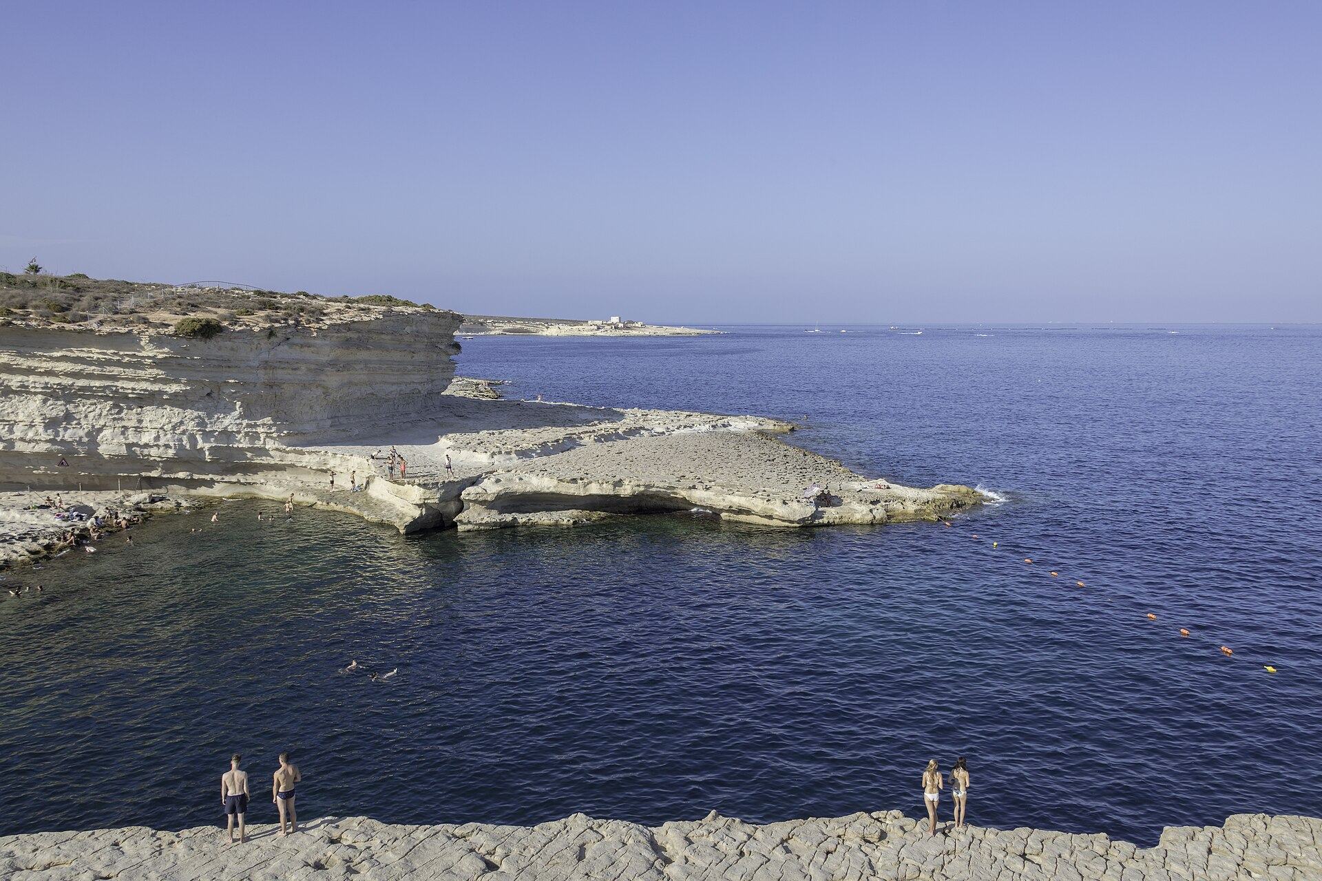 The flat pale limestone shelf at St Peter's Pool on the Delimara peninsula, with people standing at the water's edge