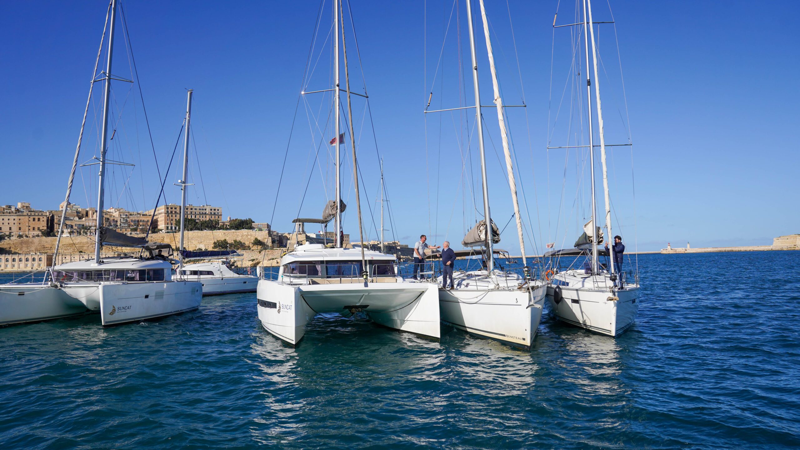 Suncat's catamarans and sailing yachts moored side by side, with the Valletta bastions and the breakwater lighthouse behind